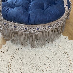 K&ouml;nnte beinhalten: Ein blaues Samtkissen mit gestepptem Design befindet sich in einem Makramee-Schaukelstuhl mit Fransenbesatz. Der Schaukelstuhl h&auml;ngt &uuml;ber einem wei&szlig;en, runden H&auml;kelteppich mit Mandala-Muster.