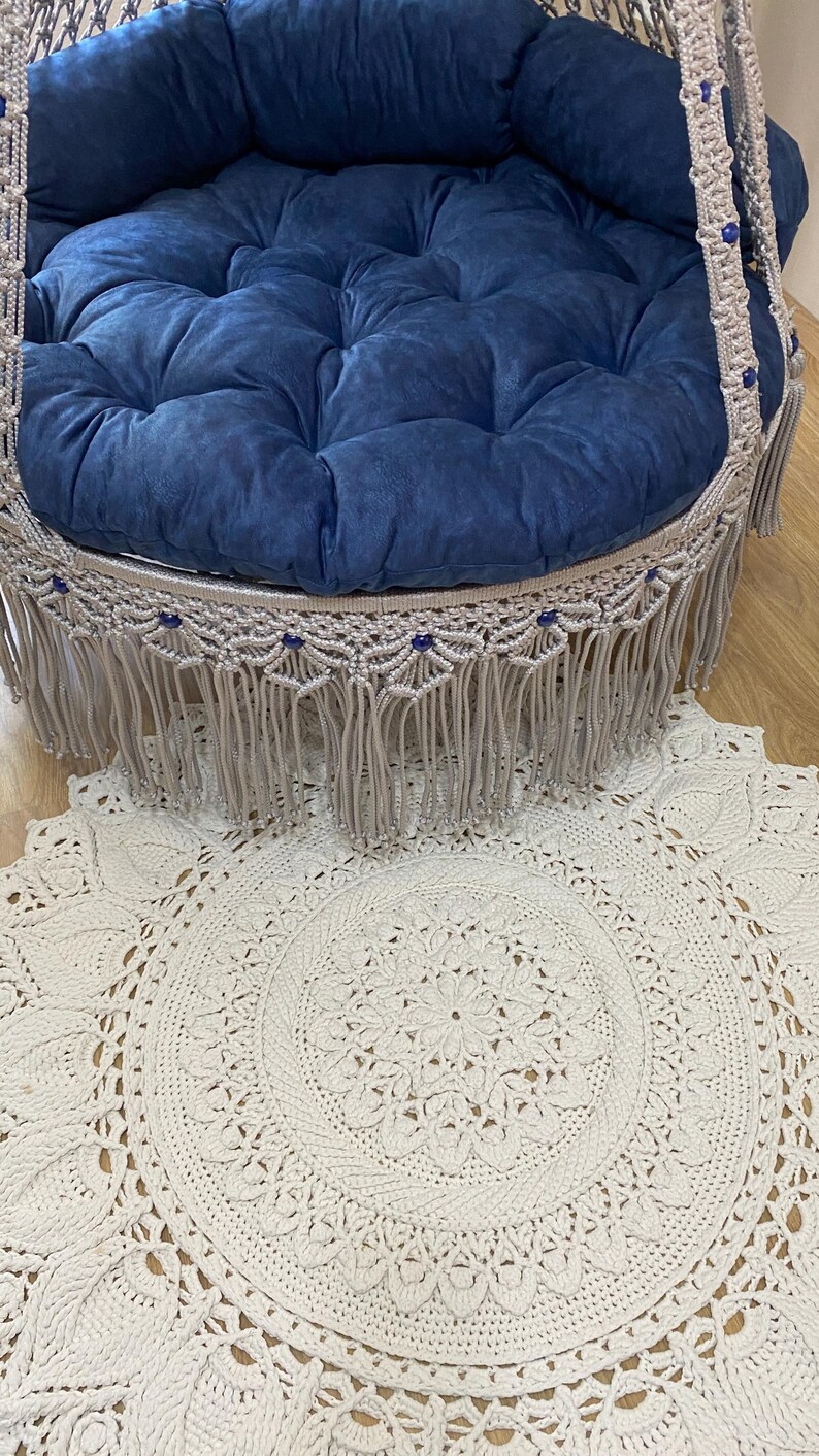 K&ouml;nnte beinhalten: Ein blaues Samtkissen mit gestepptem Design befindet sich in einem Makramee-Schaukelstuhl mit Fransenbesatz. Der Schaukelstuhl h&auml;ngt &uuml;ber einem wei&szlig;en, runden H&auml;kelteppich mit Mandala-Muster.