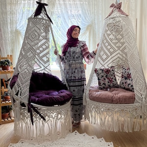 May include: Two white macrame hanging chairs with fringe, one with a purple cushion and the other with a pink cushion. The chairs are hanging from the ceiling in a room with white curtains.