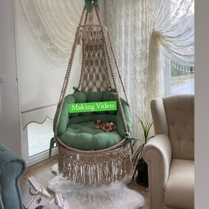 Könnte beinhalten: Ein hängender Makramee-Stuhl mit einem grünen Kissen und Fransen. Der Stuhl hängt von der Decke in einem Raum mit weißen Vorhängen.