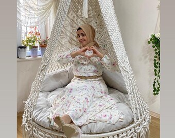 Könnte beinhalten: Ein Makramee-Hängesessel mit weißem Baldachin und rundem Kissen. Der Stuhl hat einen gefransten Boden und ist an der Decke aufgehängt. Die Person im Stuhl trägt ein Blumenkleid.