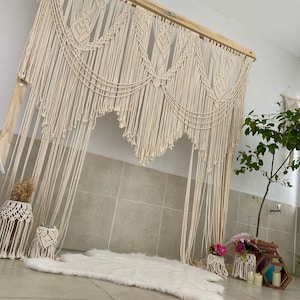 May include: A macrame wall hanging with a wooden frame, featuring intricate knotting patterns and a flowing, fringed design. The neutral color palette complements the bohemian style. Decorative macrame plant hangers and a small rug are also visible.