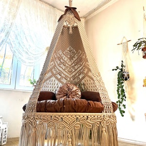 May include: A beige macrame hanging chair with a brown cushion and a pink pillow. The chair is suspended from the ceiling with a chain and has a fringe around the bottom.