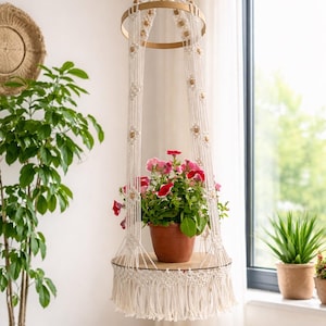 May include: A macrame plant hanger with a wooden shelf, holding a terracotta pot with vibrant pink and red flowers. The off-white macrame design features wooden beads and fringe, creating a bohemian aesthetic. The hanger is suspended from a wooden ring.