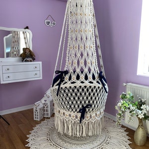 K&ouml;nnte beinhalten: Ein wei&szlig;er Makramee-H&auml;ngesessel mit einem blauen Kissen und Fransen. Der Sessel h&auml;ngt von der Decke in einem Raum mit einer wei&szlig;en Kommode und einer violetten Wand.