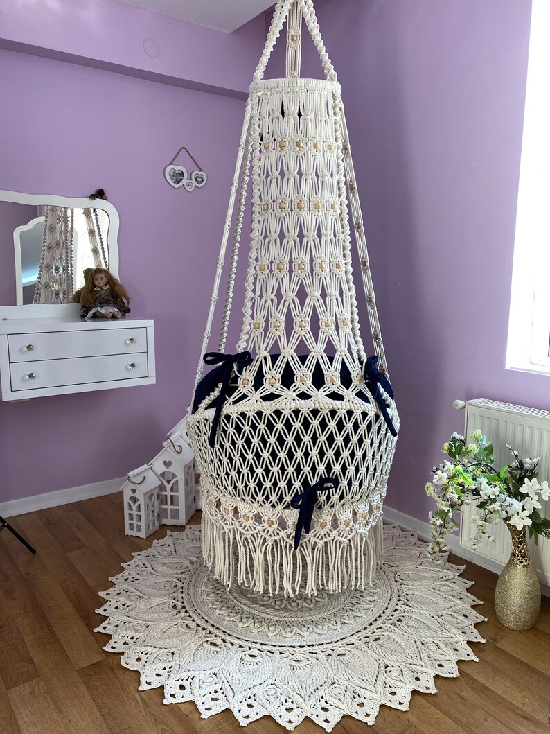 K&ouml;nnte beinhalten: Ein wei&szlig;er Makramee-H&auml;ngesessel mit einem blauen Kissen und Fransen. Der Sessel h&auml;ngt von der Decke in einem Raum mit einer wei&szlig;en Kommode und einer violetten Wand.