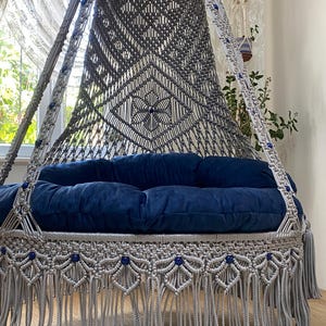K&ouml;nnte beinhalten: Ein grauer Makramee-H&auml;ngesessel mit einem blauen Kissen und blauen Perlen. Der Sessel hat einen Fransenrand und h&auml;ngt von der Decke.