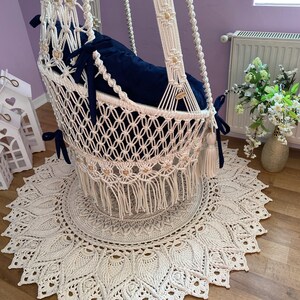 K&ouml;nnte beinhalten: Ein wei&szlig;er Makramee-H&auml;ngesessel mit einem blauen Kissen und Holzperlen. Der Sessel h&auml;ngt &uuml;ber einem wei&szlig;en Teppich mit einem runden Design.