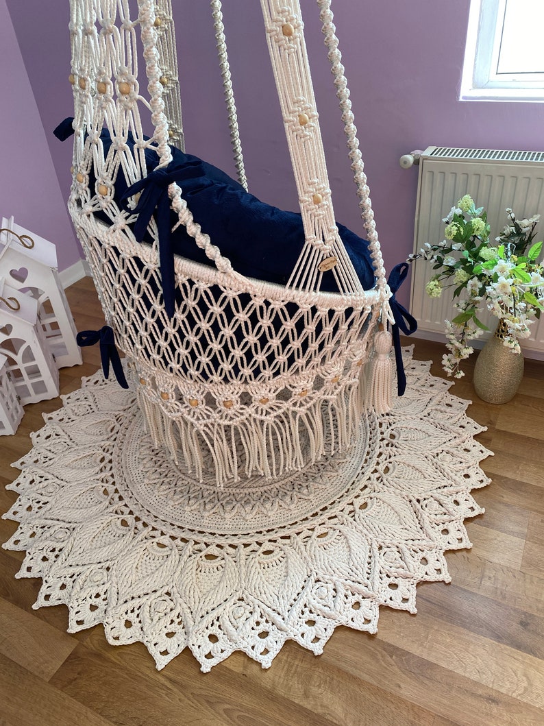 K&ouml;nnte beinhalten: Ein wei&szlig;er Makramee-H&auml;ngesessel mit einem blauen Kissen und Holzperlen. Der Sessel h&auml;ngt &uuml;ber einem wei&szlig;en Teppich mit einem runden Design.