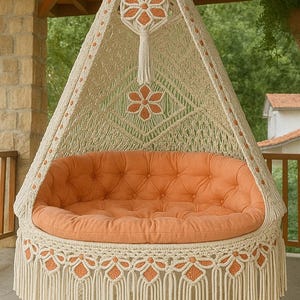 May include: A handcrafted macrame swing chair with an orange tufted cushion. The off-white chair features intricate knotwork, orange floral accents, and a fringed skirt. A decorative bow tops the hanging structure, creating a cozy outdoor seating option.