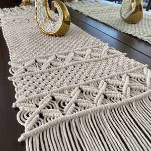 Pode incluir: Um corredor de mesa de macramé bege com um padrão geométrico e franjas. O corredor está sobre uma mesa de madeira marrom escura.