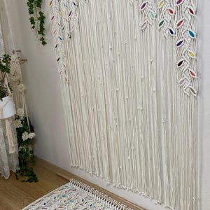 Puede incluir: Un tapiz de macram&eacute; blanco con detalles coloridos. El tapiz presenta un patr&oacute;n repetitivo de hojas y est&aacute; hecho con cord&oacute;n de algod&oacute;n grueso.