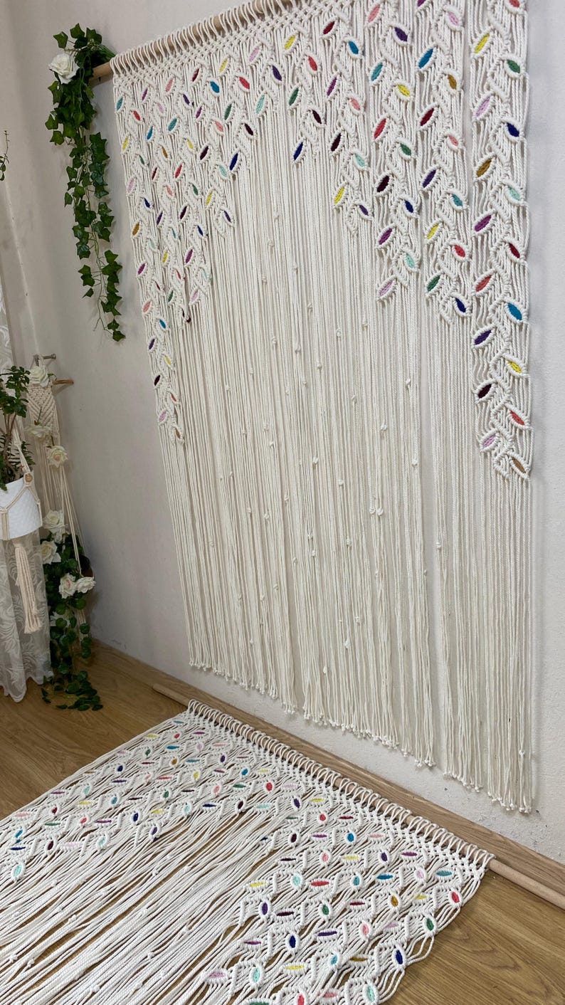 Puede incluir: Un tapiz de macram&eacute; blanco con detalles coloridos. El tapiz presenta un patr&oacute;n repetitivo de hojas y est&aacute; hecho con cord&oacute;n de algod&oacute;n grueso.