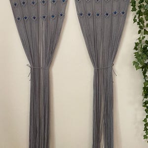 Könnte beinhalten: Grauer Makramee-Wandbehang mit blauen Perlen, mit geometrischem Muster. Der Behang besteht aus mehreren Garnsträngen und ist unten zusammengebunden.
