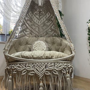 May include: A beige macrame swing with a cushion and pillow. The swing has a fringe trim and is hanging from a white ceiling. The swing is sitting on a beige crocheted rug.