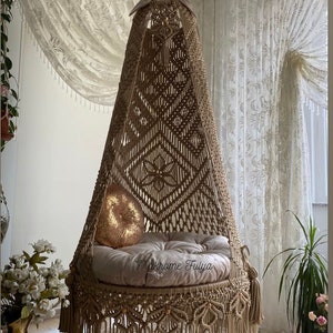 Puede incluir: Una silla colgante de macramé beige con un borde de flecos y un cojín blanco. La silla está colgada de un techo blanco y está delante de una cortina blanca transparente.