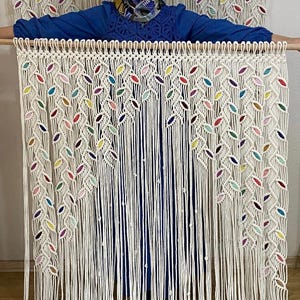 Puede incluir: Un tapiz de macram&eacute; blanco con detalles coloridos. El tapiz presenta un patr&oacute;n de hojas y est&aacute; hecho con cord&oacute;n de algod&oacute;n grueso.