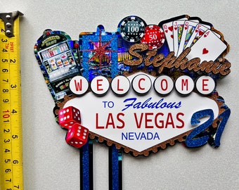May include: A Las Vegas themed cake topper with a welcome to Las Vegas sign, playing cards, dice, and a slot machine. The sign is gold and blue with red lettering. The cake topper says "Welcome to Fabulous Las Vegas Nevada" and "Stephanie 21".