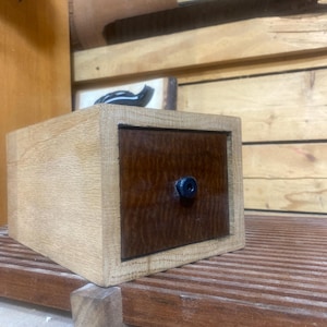 Puede incluir: Una pequeña caja de madera con un cajón. El cajón está hecho de madera oscura con un patrón de veta de madera. La caja está sentada sobre una superficie de madera.