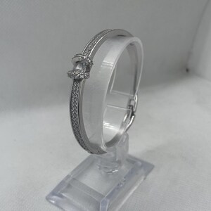 May include: A silver-colored bangle bracelet with a row of small, clear stones along the top and bottom edges. The bracelet has a knot-like design in the center and is displayed on a clear stand.