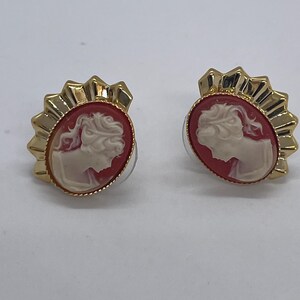 May include: A pair of gold-toned cameo stud earrings. Each earring features a red oval cameo with a white profile and is surrounded by a gold, sunburst-style frame. The earrings are set against a white background.