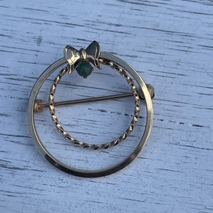 May include: A gold-toned brooch with a green stone in the center. The brooch is shaped like a circle with a smaller circle inside. The smaller circle is made of twisted wire.