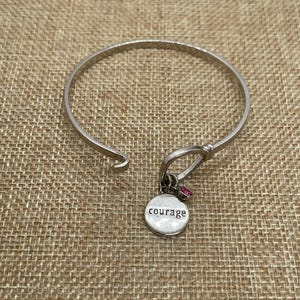 May include: Silver bangle bracelet with a charm that says "courage" and two pink rhinestones.