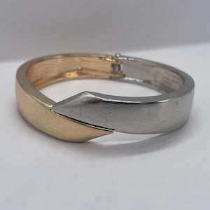 May include: A two-tone hinged bracelet. The bracelet features a gold-colored section and a silver-colored section that meet in a geometric design. The bracelet is a simple, yet elegant piece of jewelry.