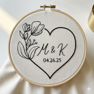 May include: Embroidered hoop art with a black heart and floral design. The initials "M & K" and the date "04.26.25" are stitched inside the heart. The embroidery is set in a wooden hoop.