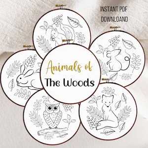 May include: Five embroidery hoop patterns featuring woodland animals: a squirrel, a mouse, a rabbit, an owl, and a fox. The hoops are white with black outlines of the animals and leaves. The text "Animals of The Woods" is in gold lettering.