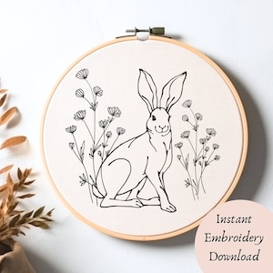 May include: Black and white line art embroidery pattern of a rabbit sitting in a field of flowers. The design is on a white fabric background in a wooden embroidery hoop.