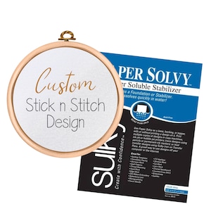 Op de afbeelding: Een houten borduurring met de woorden "Custom Stick n Stitch Design". Een blauw en zwart pakket "PER SOLVY" in water oplosbare stabilisator ligt naast de ring.