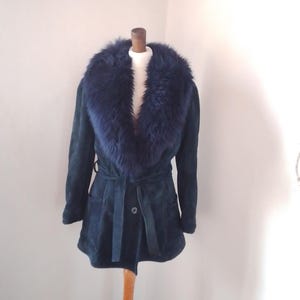 May include: A dark blue suede coat with a faux fur collar. The coat has a belted waist, two front pockets, and a button closure. The coat is displayed on a mannequin.