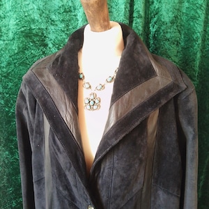 Op de afbeelding: Een zwarte suède jas met leren accenten en een turquoise knoop. De jas heeft een brede kraag en een decoratieve ketting met turkooizen stenen. De jas wordt gepresenteerd op een mannequin tegen een groene achtergrond.