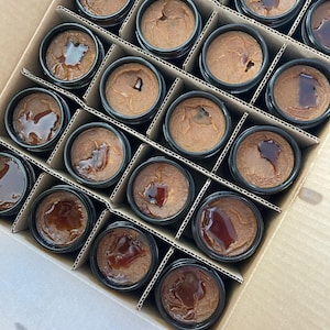 May include: A cardboard box filled with small glass jars containing a brown substance. The jars are arranged in rows and columns, and the box is open to reveal the contents.