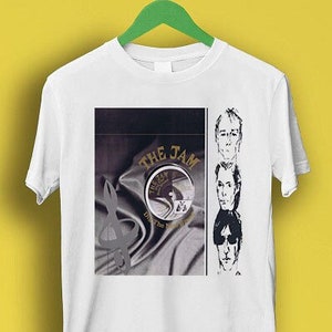 以下が含まれることがあります： 白地に、バンド「ザ・ジャム」の白黒画像がプリントされたTシャツ。画像にはバンド名、レコード、バンドメンバー3人の顔が写っている。