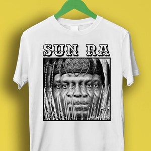 Op de afbeelding: Wit T-shirt met een zwart-witte afbeelding van Sun Ra, een muzikant en componist, die een kroon en traditionele Afrikaanse kleding draagt. De tekst "SUN RA" is in zwart boven de afbeelding gedrukt.