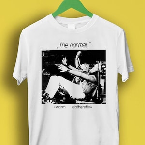 Puede incluir: Camiseta blanca con una foto en blanco y negro de una persona en un coche. El texto "the normal" y "warm leatherette" está impreso en la camiseta.