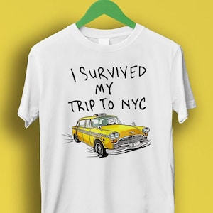 Może przedstawiać: Biała koszulka z napisem "I survived my trip to NYC" i rysunkiem żółtej taksówki.