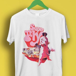May include: White t-shirt featuring a pink and red graphic of a man in a white suit holding a gun, standing next to a gold car. The graphic also includes the text "Super Fly" in pink.