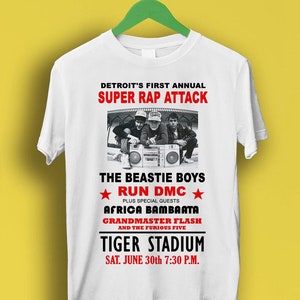 May include: A white t-shirt featuring a black and white graphic of a concert poster for Detroit's First Annual Super Rap Attack. The poster features The Beastie Boys, Run DMC, Afrika Bambaata, Grandmaster Flash and The Furious Five. The concert is at Tiger Stadium on June 30th at 7:30 PM.
