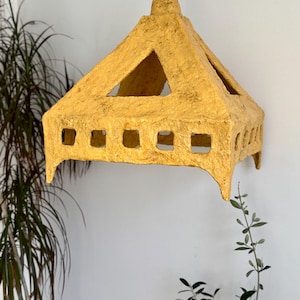 May include: A handmade, yellow paper pendant light fixture. The light has a geometric design with a pyramid-shaped top and rectangular cutouts along the sides. The light hangs from a brown cord, and is mounted on a white wall.