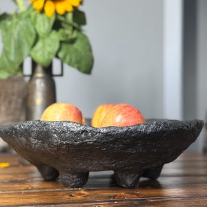 May include: A black, textured, footed bowl holds two red apples. The bowl is on a wooden surface, with a sunflower in a vase in the background.