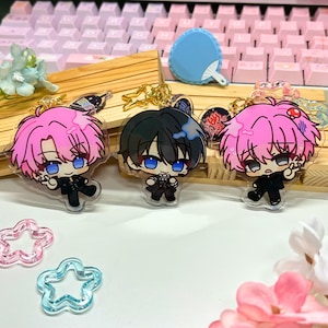 May include: Three anime-style acrylic keychains featuring characters with pink and black hair, blue eyes, and black outfits. The keychains are on a wooden display with a pink keyboard in the background. Additional accessories include a blue fan and star-shaped charms.