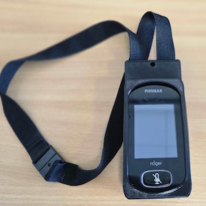 Puede incluir: Un dispositivo de audición negro con una pantalla que dice "roger" y un icono de micrófono. El dispositivo está conectado a un cordón negro.