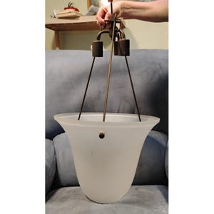 May include: A frosted glass pendant light fixture with a bell-shaped shade. The fixture has a bronze-colored metal frame with three arms and a central rod. The shade is suspended by thin wires. The light is held by a person's hand.