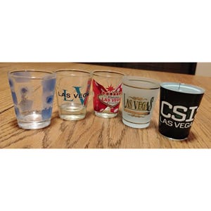 May include: Five clear shot glasses with various designs and the words "Las Vegas" and "CSI Las Vegas" printed on them. The shot glasses are on a wooden surface.