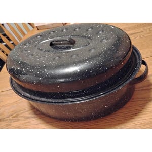 May include: A black speckled roasting pan with a matching lid. The lid has a handle and small holes. The pan has two handles on the sides. The pan is sitting on a wooden surface.