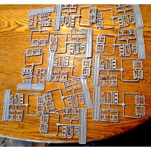 May include: A collection of gray plastic model kit parts, still attached to their sprues, scattered on a wooden surface. The parts appear to be for a building or vehicle, with various shapes and sizes. The image is taken from a top-down perspective.