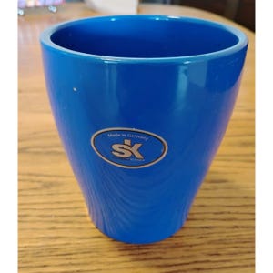 May include: A vibrant blue ceramic planter with a glossy finish. The planter has a slightly tapered shape and features a gold-toned sticker that reads "Made in Germany" and the initials "SK". The planter is empty and ready for use.